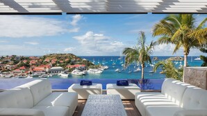 Una terraza con sofás blancos y una mesa de vidrio con vistas a un puerto deportivo con barcos y una ciudad costera.