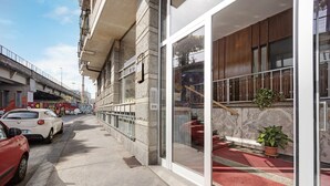 Un bâtiment dont l’entrée est recouverte d’un tapis rouge, qui possède un escalier et un balcon agrémenté d’un pot de fleurs.