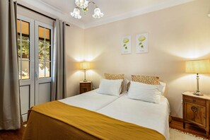 Ein Doppelbett mit weißer und brauner Bettdecke, zwei Nachttische mit Lampen, ein Fenster mit Vorhängen und zwei an der Wand befestigte Blumenbilder.