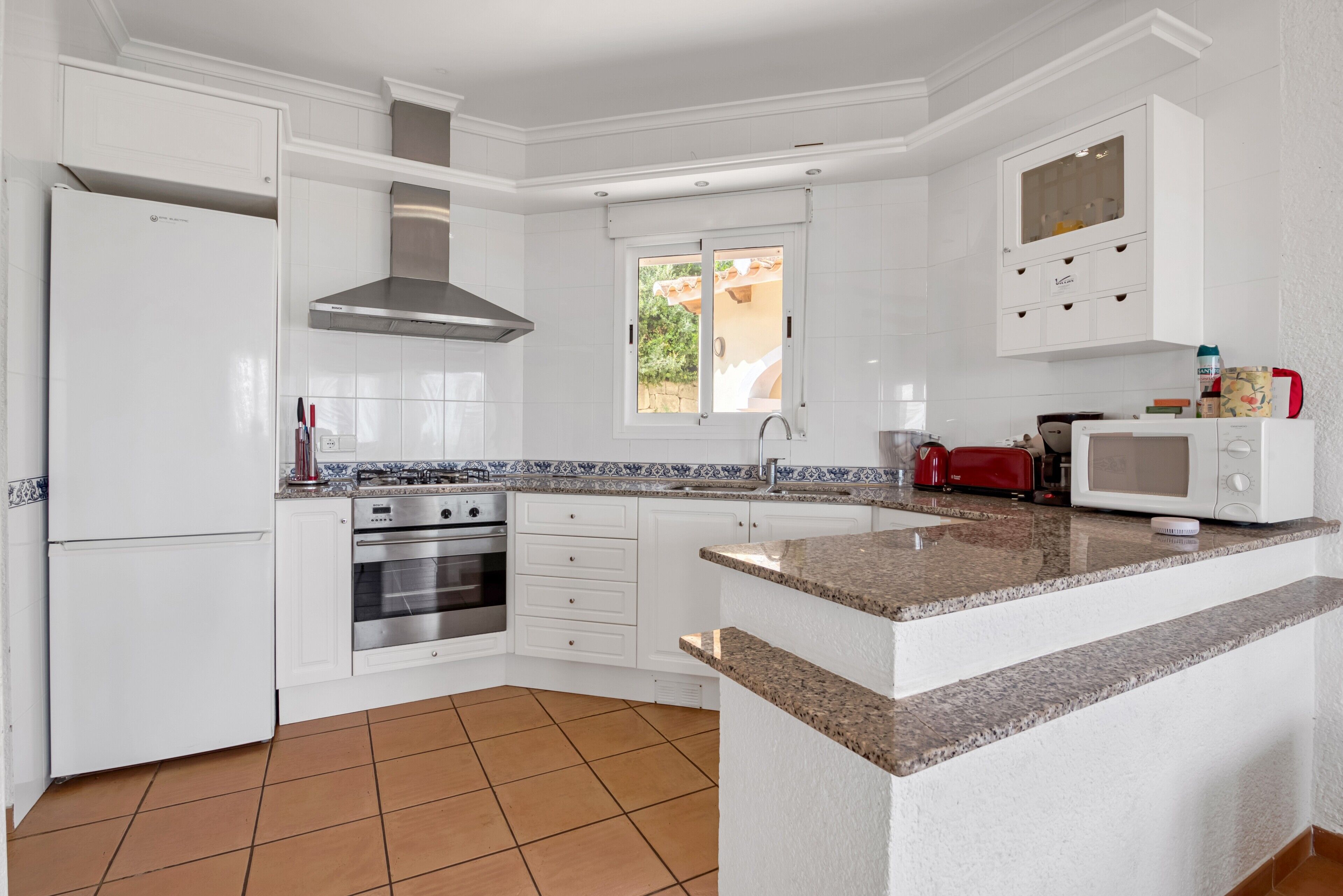 Una cocina moderna con armarios blancos, estufa de acero inoxidable, horno empotrado, microondas y un ventanal que proporciona luz natural.