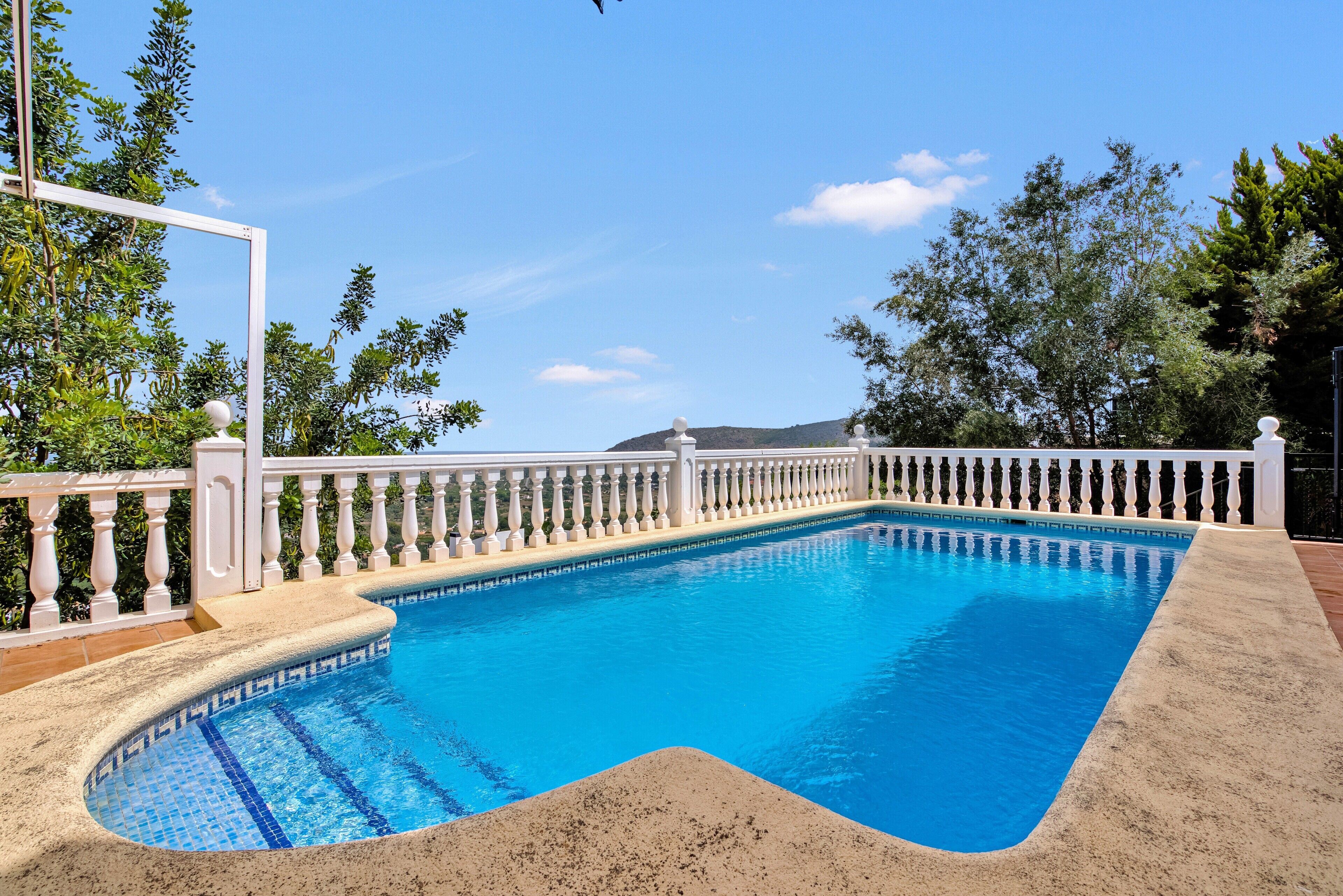 Una piscina rectangular con barandilla blanca, rodeada de follaje y un cielo despejado.