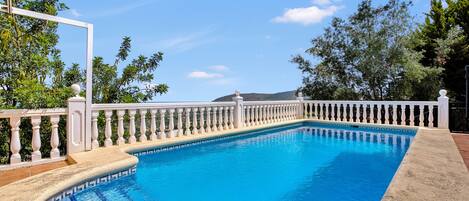 Una piscina rectangular con barandilla blanca, rodeada de follaje y un cielo despejado.