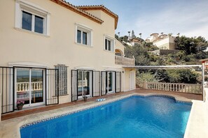 Uma piscina com borda em azulejo, uma varanda com grade e vista para uma encosta com edifícios.