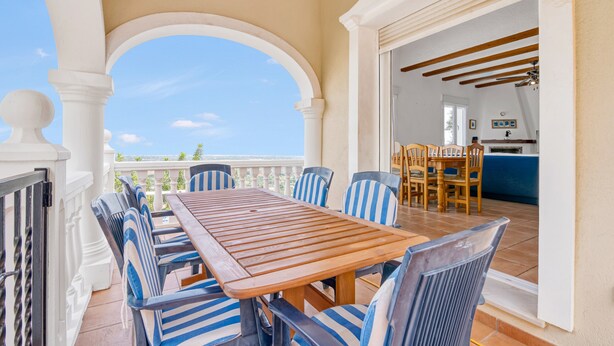 Un espace extérieur abrité, aménagé pour prendre les repas, avec une table en bois et des chaises à rayures, une porte ouverte donnant sur une cuisine et une vue sur le ciel.