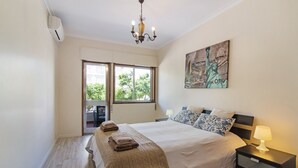 Quarto com cama, mesa de cabeceira, abajur, pintura da Estátua da Liberdade e janela com vista para vegetação.