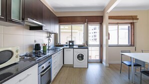 Uma cozinha compacta com fogão, micro-ondas e máquina de lavar, uma área de refeições com cadeiras, e vista pela porta.