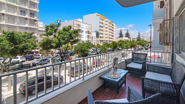 Uma varanda com mobiliário de vime, vista para uma rua movimentada e edifícios ao fundo.