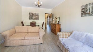 Uma sala de estar com um sofá bege, um sofá branco, um candelabro e uma área de jantar ao fundo.