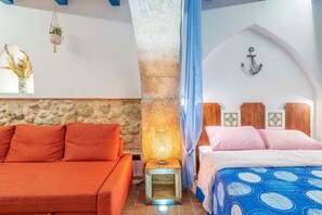 A room with a stone wall, a bed with a blue patterned bedspread, an orange sofa, a small table with a lamp, and a wall-mounted light fixture.