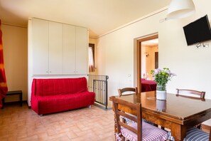 A room with a red sofa, a wooden dining table with chairs, a TV mounted on the wall, and a small vase with flowers on the table. - Holiday Home 'Calla' with Shared Pool, Wi-Fi and Air Conditioning (montecarlo)