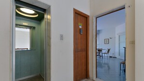 Un couloir intérieur avec une porte en bois, un ascenseur en verre et un coin repas comprenant des chaises et une table.