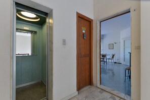Un couloir intérieur avec une porte en bois, un ascenseur en verre et un coin repas comprenant des chaises et une table.