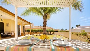 An outdoor dining area with a table set for two, a palm tree, and a view of a building in the background. - Villa 'Retiro Praia' with Mountain View, Private Terrace and Wi-Fi (Campo de Baixo)
