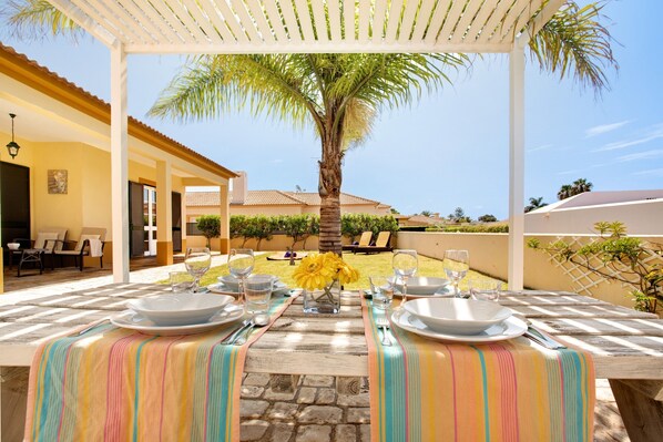 An outdoor dining area with a table set for two, a palm tree, and a view of a building in the background. - Villa 'Retiro Praia' with Mountain View, Private Terrace and Wi-Fi (Campo de Baixo)