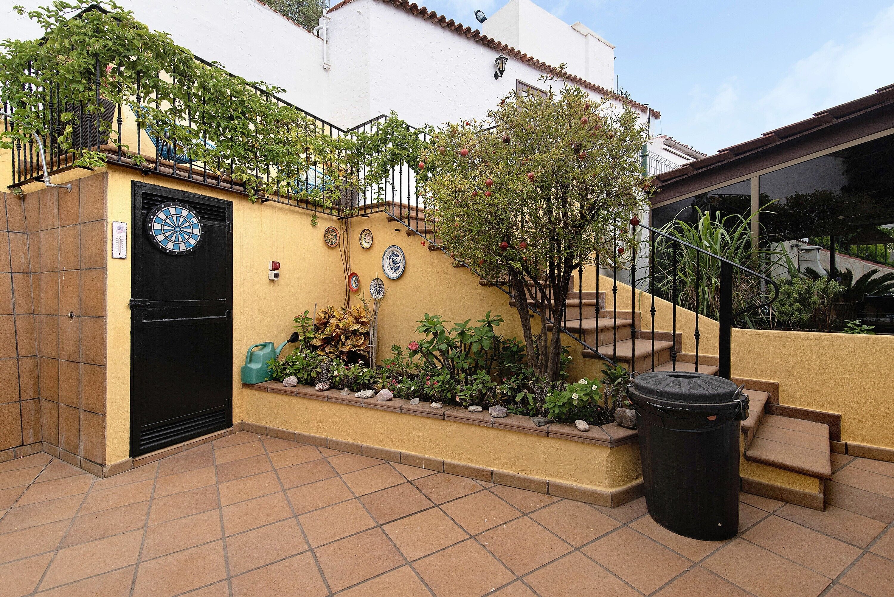 Pátio com uma porta preta, um contentor de lixo, uma escadaria e uma parede com pratos decorativos.