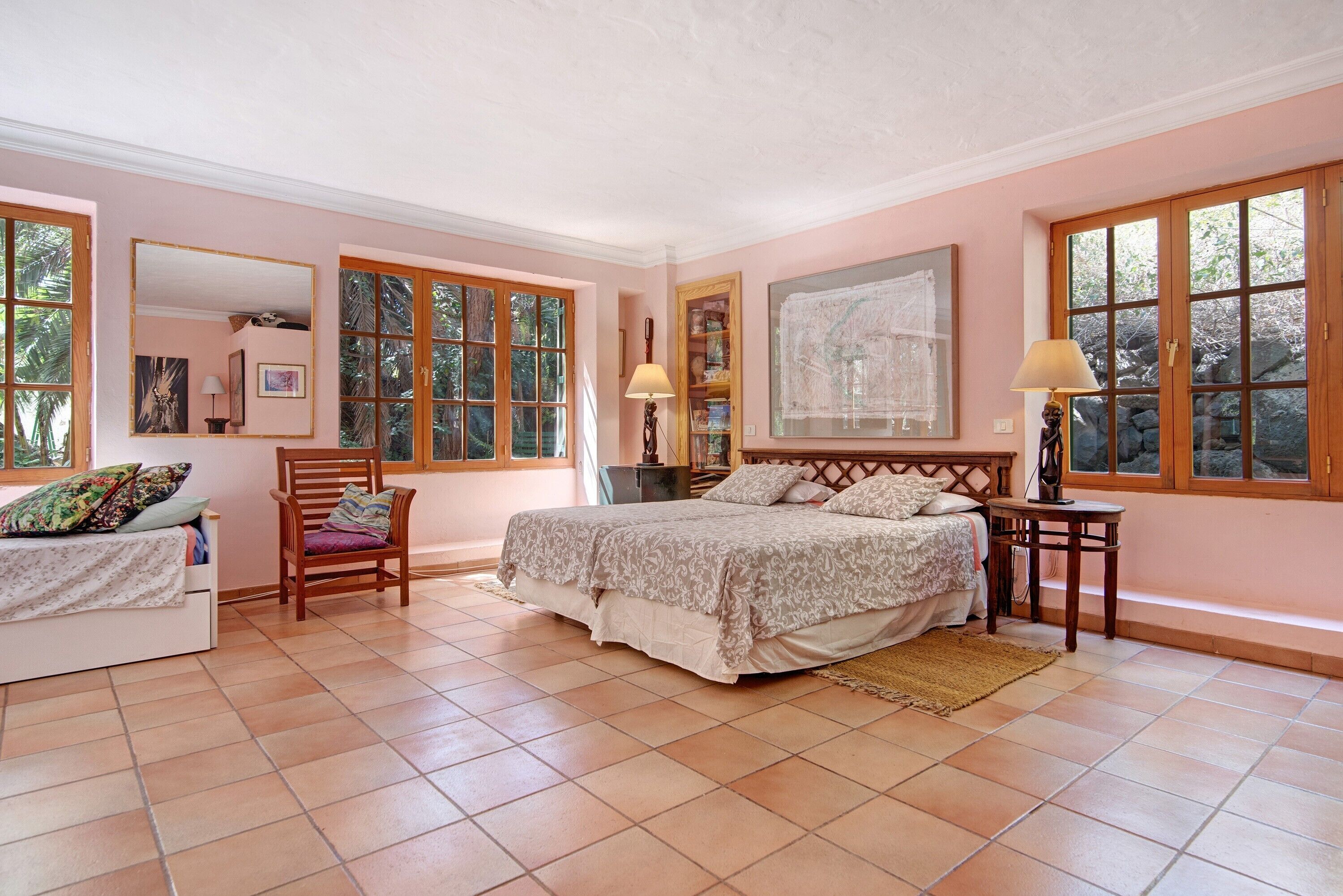 A bedroom with a bed, a chair, a bedside table, a lamp, a mirror, and large windows.