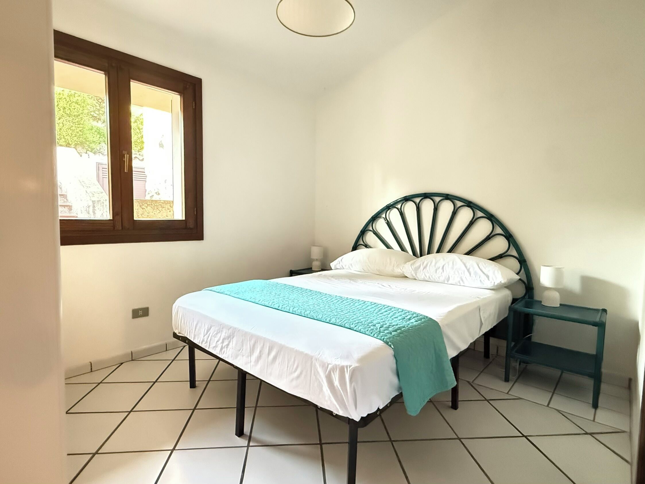 Een slaapkamer met een bed, nachtkastje, raam en betegelde vloer.