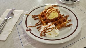 Lautasella vohveleita, päälle kermavaahtoa ja karamellikastiketta, pöydällä kahvikuppi ja aterimet.