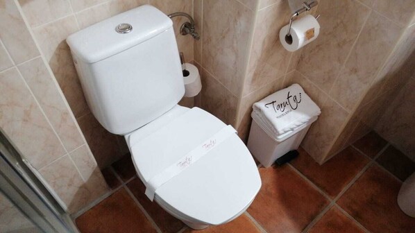 Des toilettes blanches, avec un porte-serviettes en papier et une poubelle, dans une salle de bain carrelée de carreaux beiges.