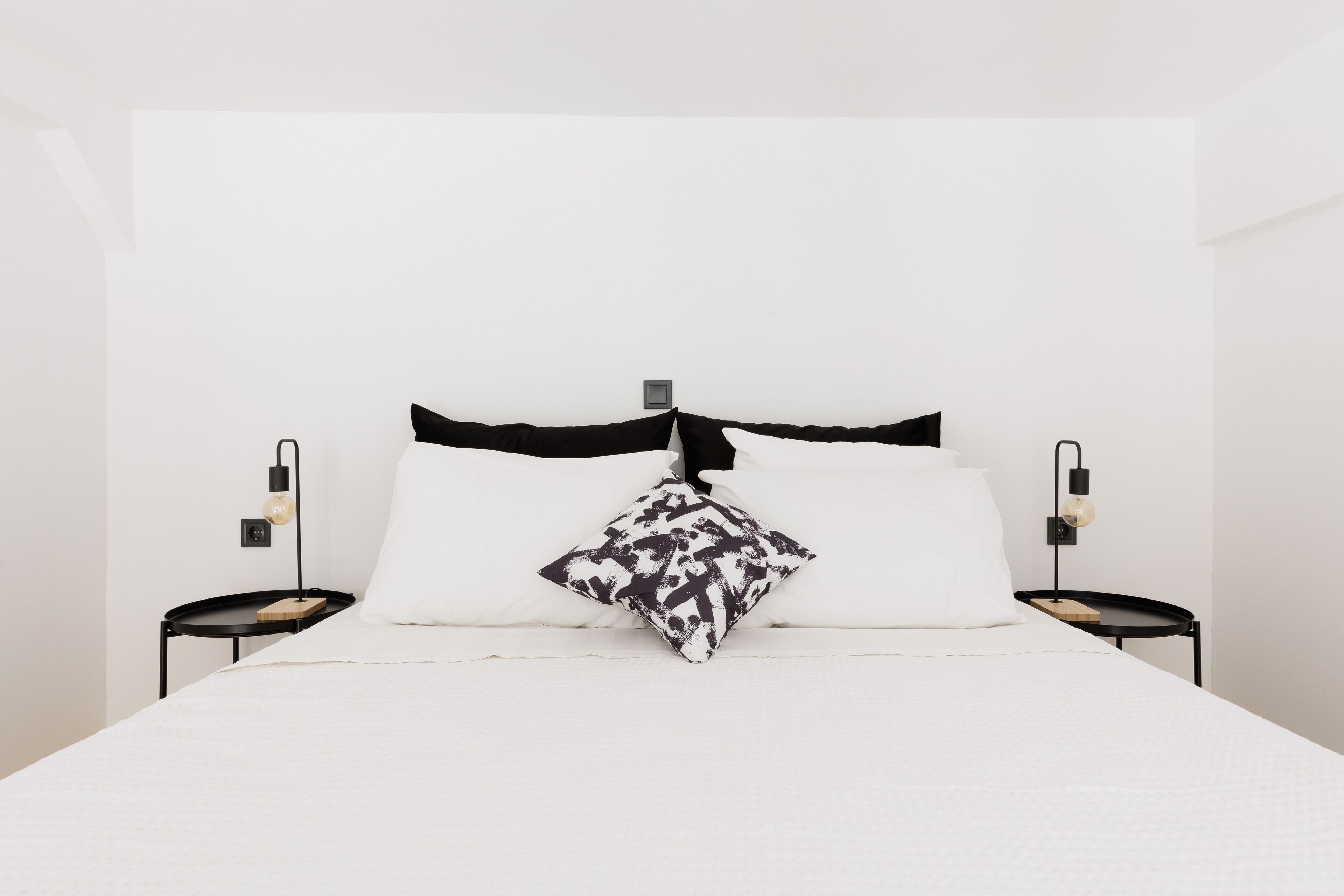 Cama bem arrumada com roupa de cama branca e almofadas de cabeceira pretas, acompanhada por duas mesinhas de cabeceira pretas com luminárias.