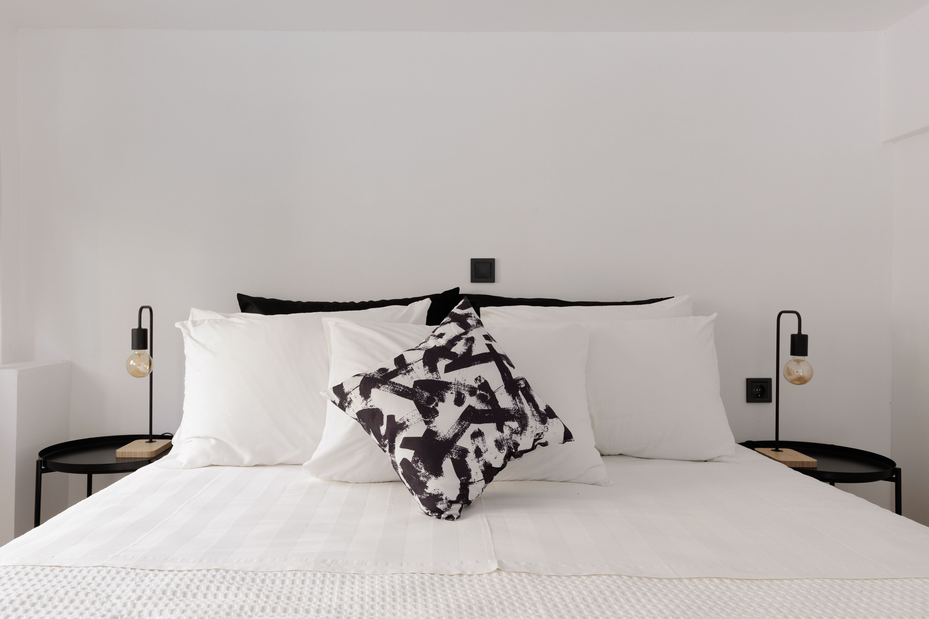 A neatly made bed with white bedding and a black and white patterned pillow. There are two bedside tables with lamps, one on each side of the bed.