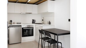 Een compacte keuken met witte kastjes, een inbouwoven, een vaatwasser en een kleine eettafel met twee stoelen.