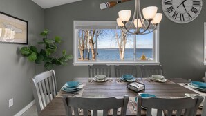 A dining room with a wooden table set for six, a large wall clock, and a view of the sea through a window. - Lakefront home, large firepit patio, Game room! (Brewerton)