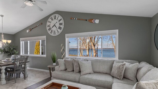 A living room with a gray sofa, a dining table with chairs, a wooden coffee table, a large wall clock, and a window with a view of trees and a body of water. - Lakefront home, large firepit patio, Game room! (Brewerton)