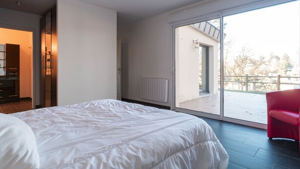 Une chambre avec un grand lit, une porte-fenêtre coulissante donnant sur un balcon et un fauteuil rouge.