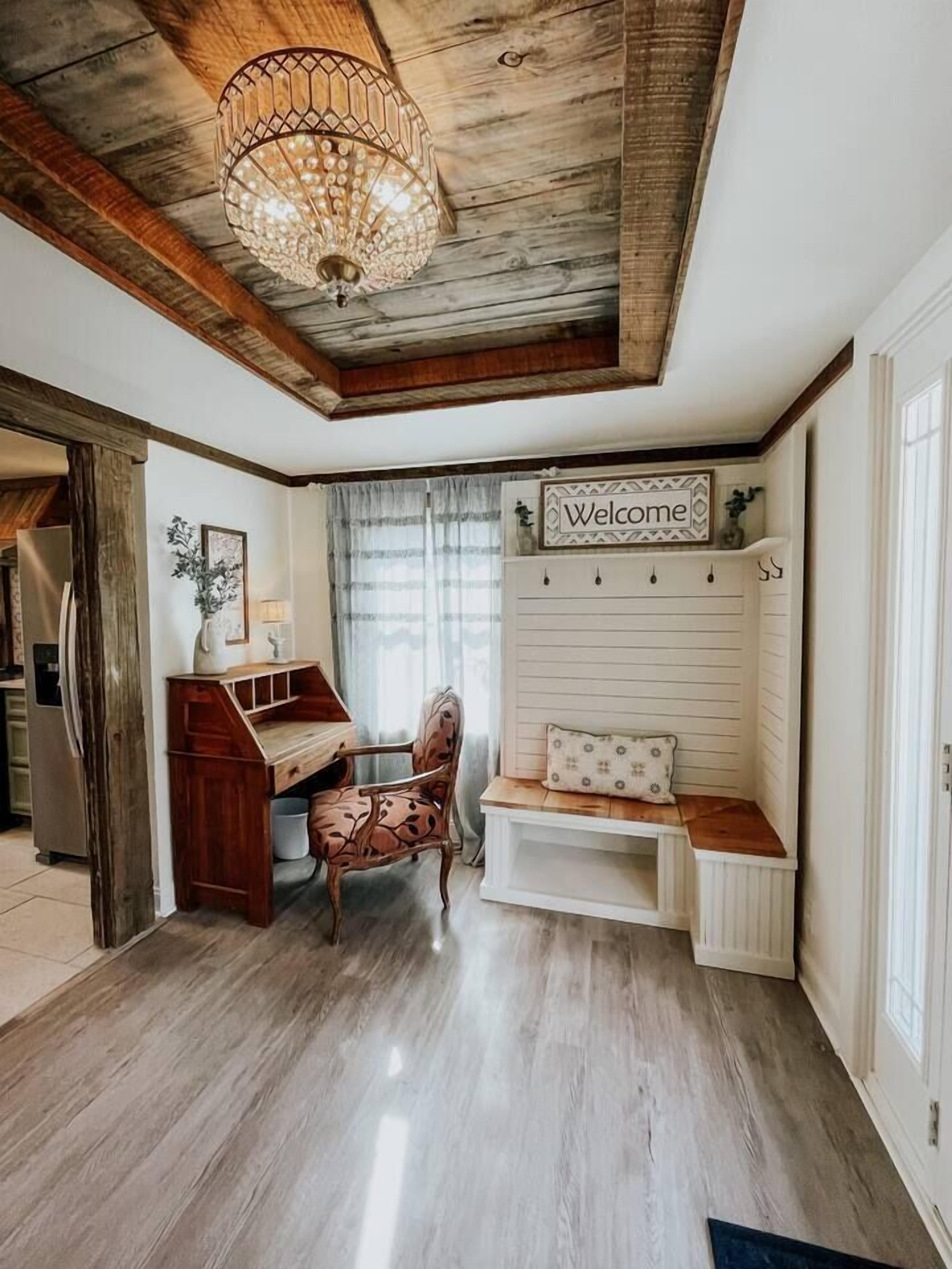 Ein gemütliches Zimmer mit Holzbalkendecke, einem Zierkronleuchter, einem 'Welcome'-Schild, einem Holztisch, einem Stuhl und einer Bank mit Kissen.