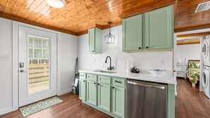 A kitchen with green cabinets, a stainless steel dishwasher, and a window with blinds. - Cozy Cottage in the Heart of Gulf Shores (Gulf Shores)