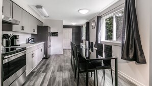 Una cocina moderna con electrodomésticos de acero inoxidable, un comedor con mesa de vidrio y sillas negras, y una ventana con cortinas oscuras.