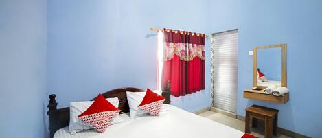 Una cama bien hecha con almohadas rojas y blancas, un cabecero de madera, un taburete de madera pequeño, un espejo sobre una repisa con toallas, y una ventana con cortinas rojas.