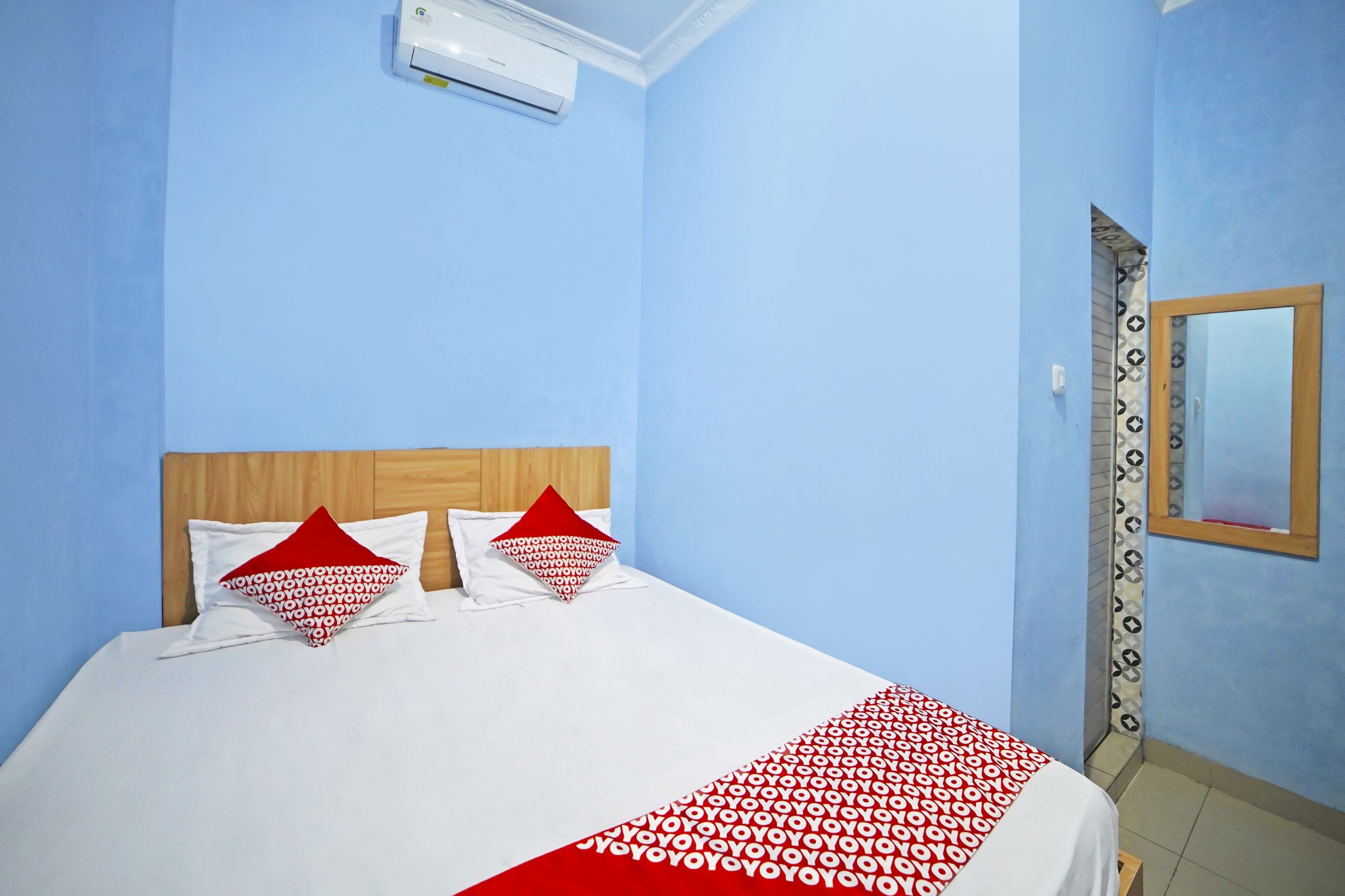Ein Hotelzimmer mit einem Bett, zwei Kissen, einer roten und weißen Decke mit Muster sowie einem Holz-Kopfende und einer Wand-Klimaanlage.