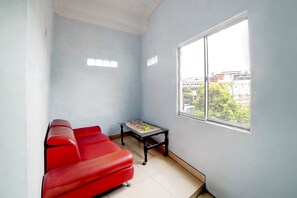 A room with a red sofa, a small table with a lace tablecloth, and a window with a view of buildings and trees. - OYO 90922 Gurusinga Guesthouse (Medan)