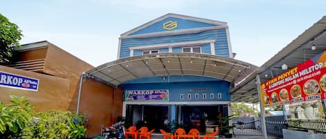 A building with a blue facade and a covered entrance. There are orange chairs outside, and a motorcycle parked nearby.