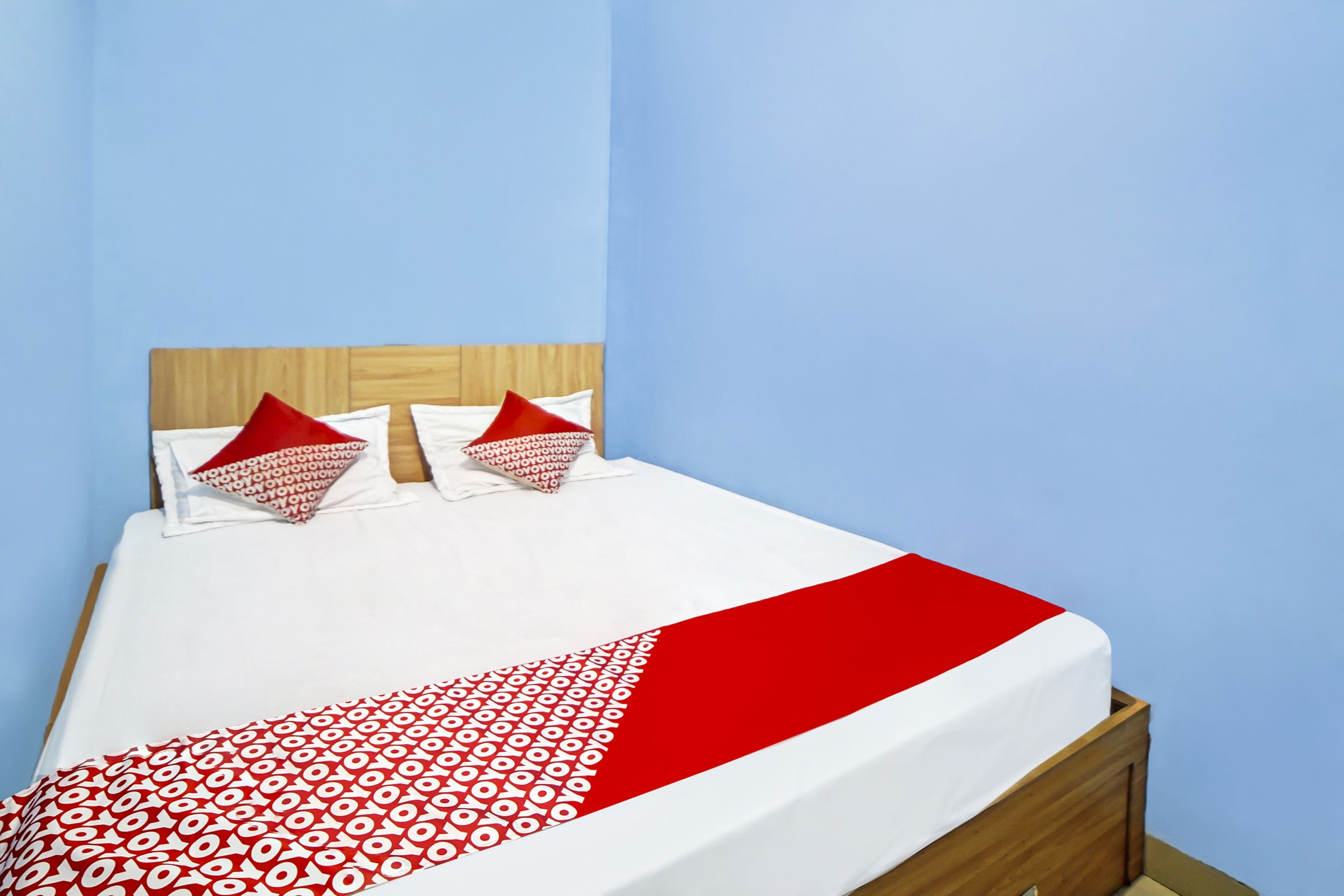 A bed with a white and red patterned bedspread, two red pillows, and a wooden headboard in a room with light blue walls.