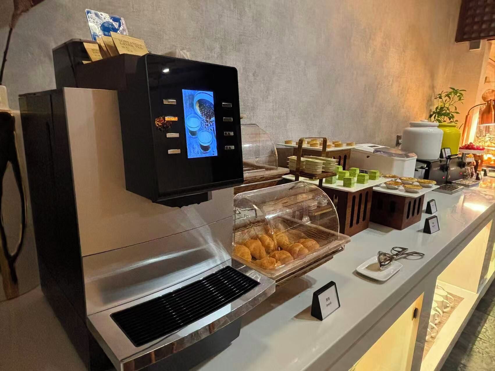 A self-service coffee station with a digital display, a variety of pastries, and a bread display.
