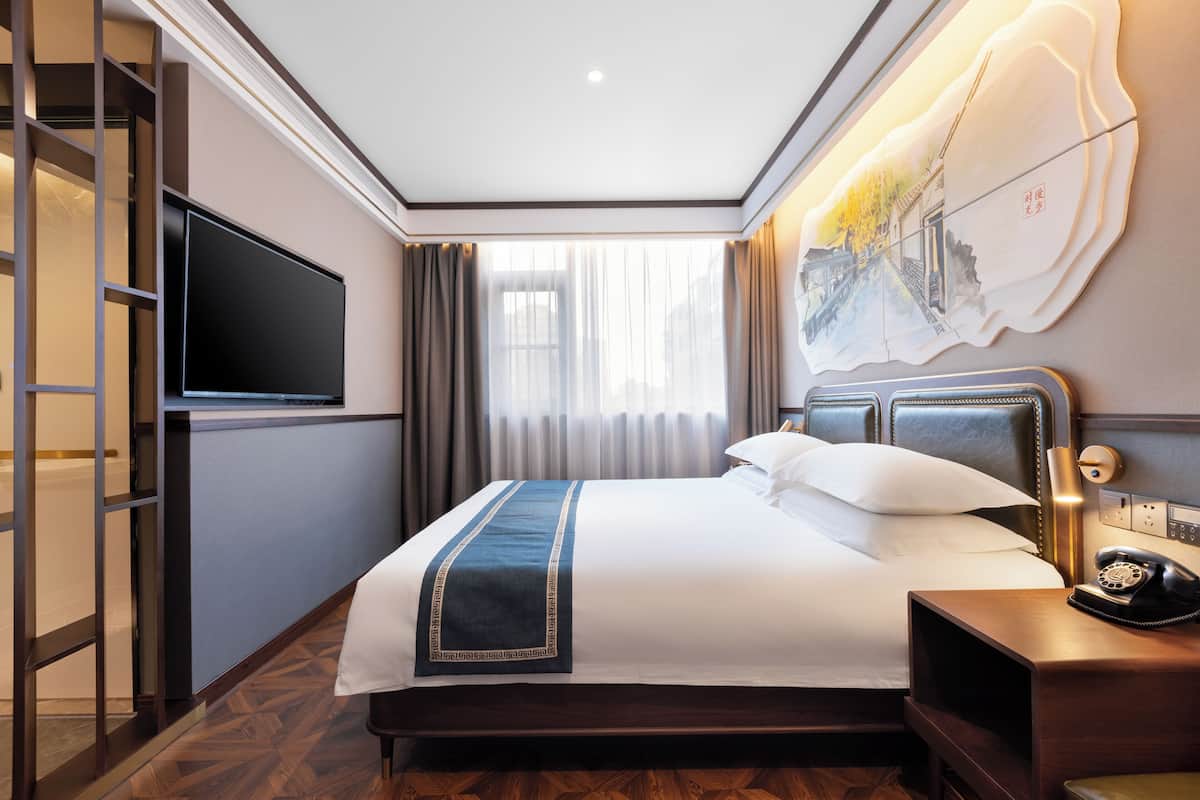 A hotel room with a bed, a TV, a bedside table with a phone, and a painting on the wall.