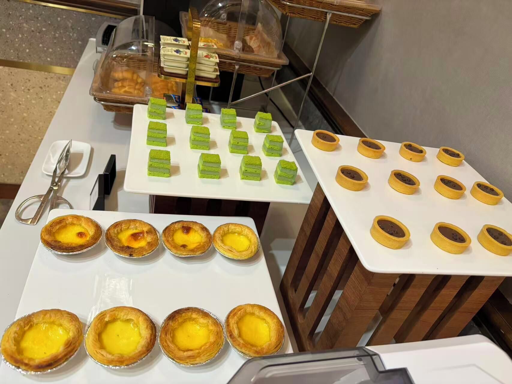 A buffet table with various pastries and desserts, including green layered cakes, yellow custard tarts, and chocolate-filled tarts.
