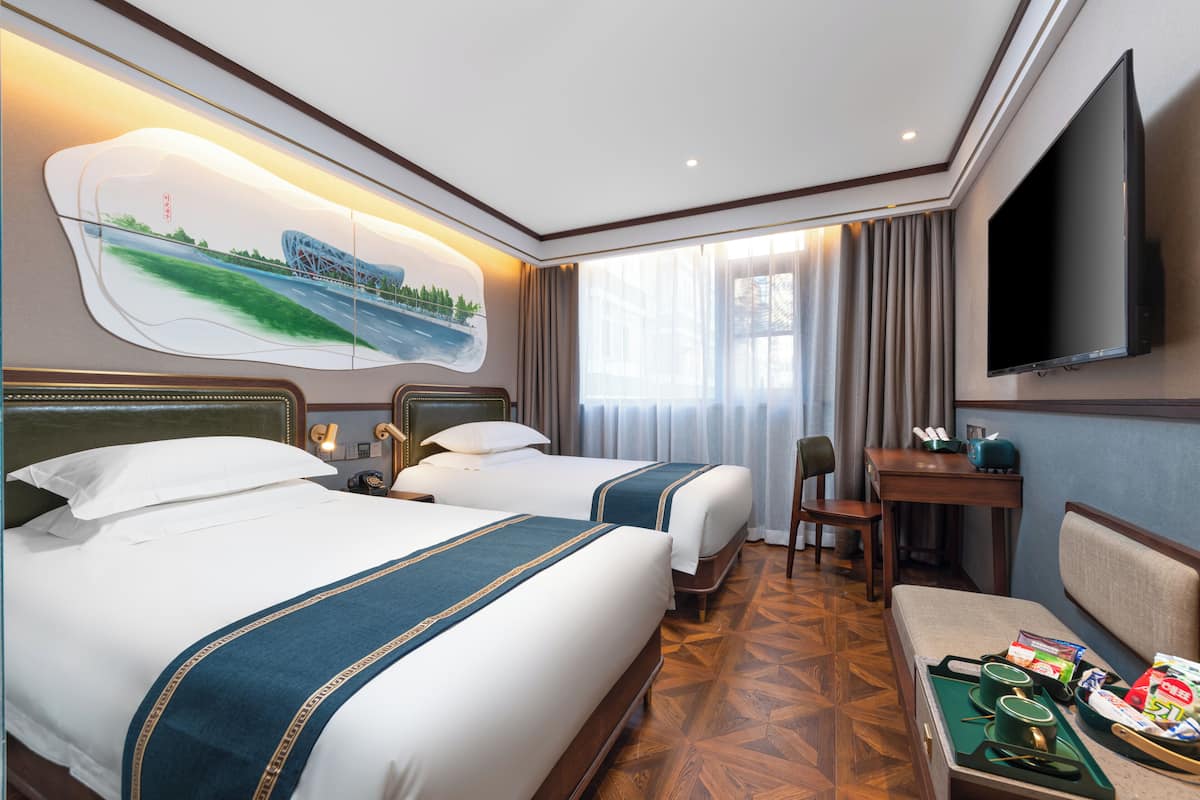 A hotel room with two beds, a desk, a chair, a TV, and a painting on the wall.