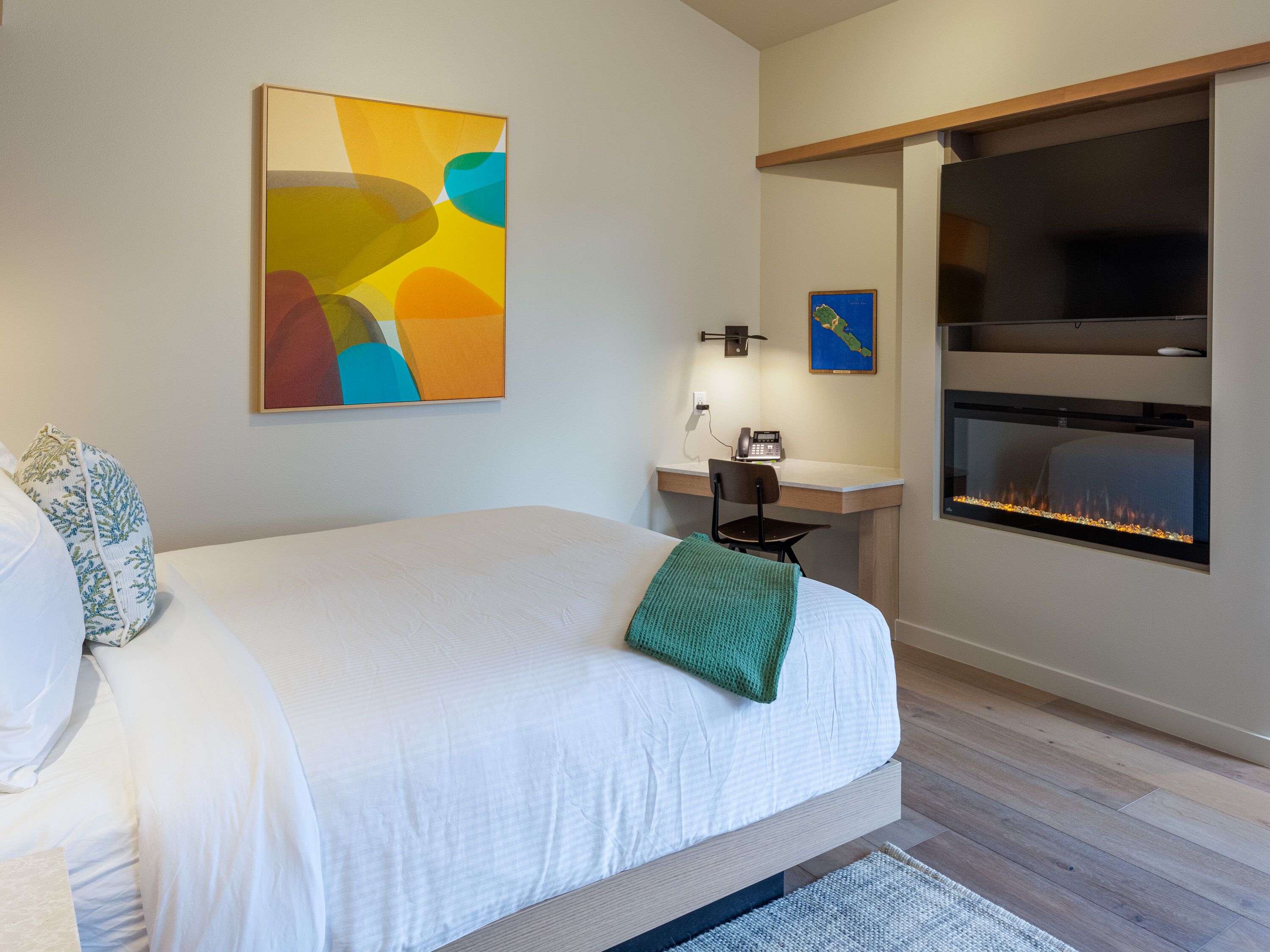 A modern hotel room with a large bed, a desk, a small chair, a wall-mounted phone, a map, and a fireplace.
