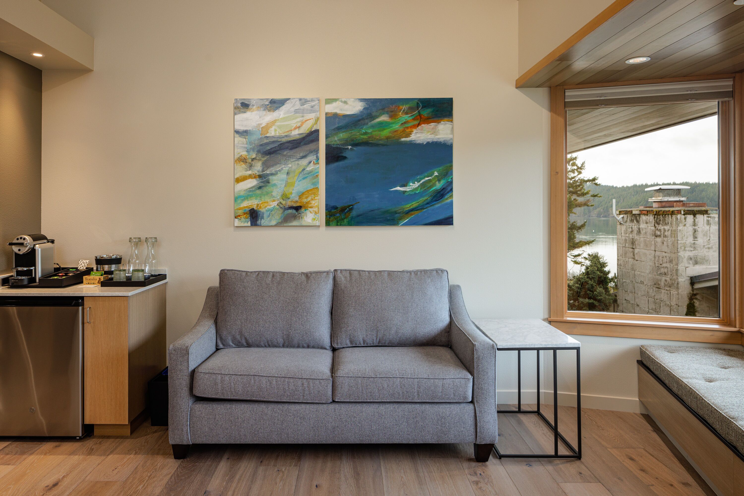 A modern living room with a grey sofa, abstract wall art, a wooden dining table, and a view of a lake through a large window.