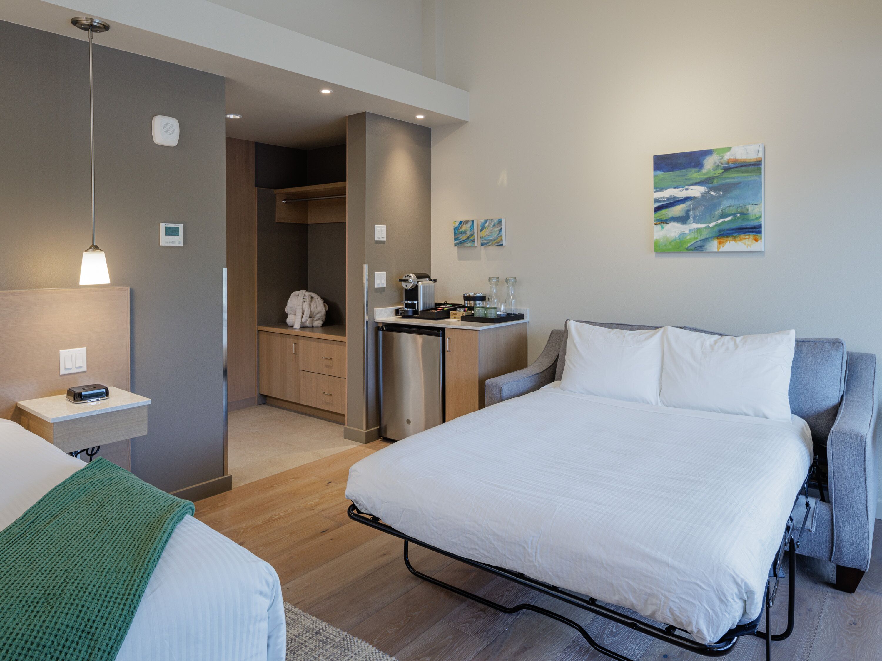 A modern hotel room with a bed, bedside table, and a kitchenette area.