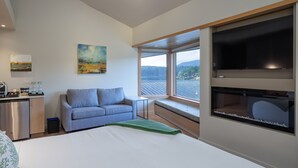 Habitación de hotel moderna con cama, sofá, zona de comedor con pequeña cocina, y un ventanal con vistas al exterior.