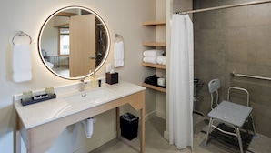 A modern bathroom with a round mirror, a sink, a towel rack, a shower area, and a small chair. - Yellow - Comfortably Accessible (Eastsound)
