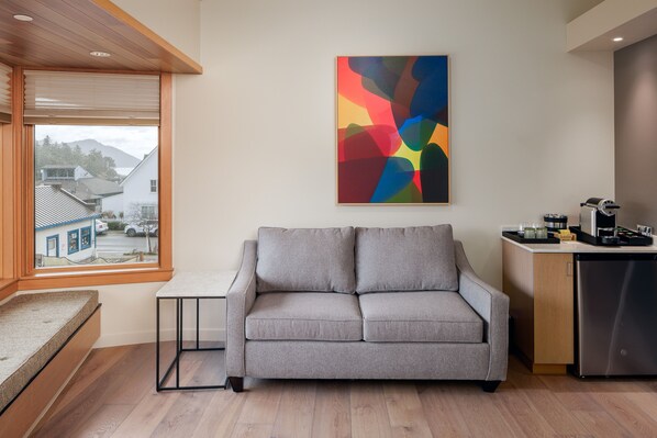 A modern living room with a grey sofa, a small table, and a kitchen area with a refrigerator.