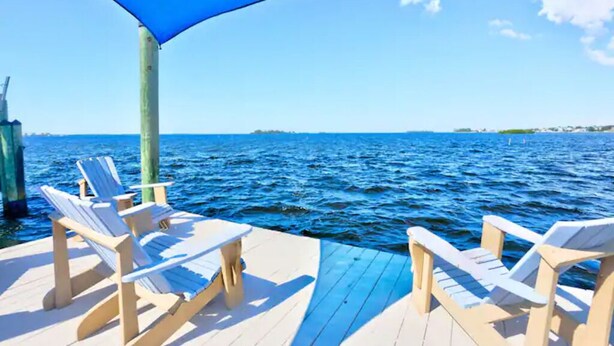 Una terraza de madera con dos sillones blancos y una mesa, con vistas a un cuerpo de agua y una orilla distante.