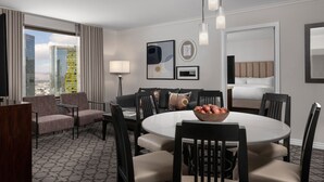 A hotel room with a dining area, a sofa, a TV, and a large window with a city view.
