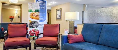 Un vestíbulo de hotel con dos sillones rojos, un sofá azul, una mesita con una lámpara y un cartel que anuncia el desayuno.